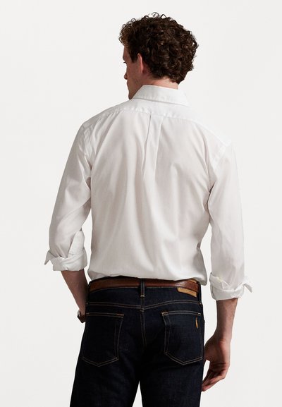 Vīrietis ar cirtainiem matiem, ģērbies baltā garu piedurkņu krekla, atloku piedurknes, un tumši zilos džinsos, redzams no aizmugures.