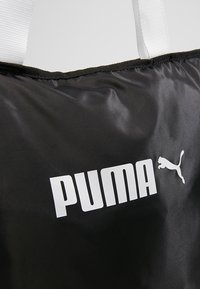 Sac Puma noir avec texte de logo blanc et icône de chat puma, avec des poignées en tissu blanc sur un fond uni.