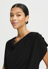 Top nero e testurizzato con scollo drappeggiato e maniche morbide, caratterizzato da un leggero luccichio. Orecchini a cerchio dorati come dettaglio accessorio notevole.