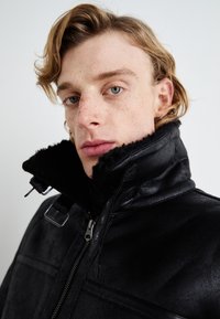 Black leather jacket with a high collar lined in black faux fur, featuring a front zipper and adjustable hardware details.