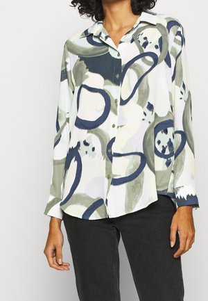 Modello che indossa una camicia con bottoni con motivi astratti verdi e blu, abbinata a pantaloni scuri, che sorride leggermente su uno sfondo chiaro.