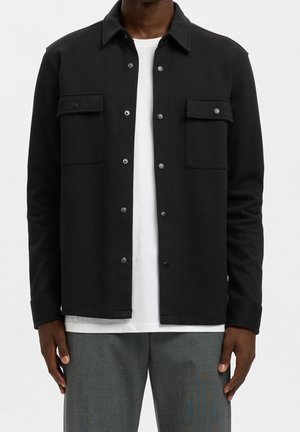 Modèle portant une veste noire à boutons sur le devant avec deux poches poitrine à rabat sur un T-shirt blanc et un pantalon gris.