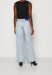 Kvinna i ljus tvättade raka jeans med bakfickor och en blå "Karl Lagerfeld Jeans"-etikett, svart ärmlös topp och svarta klackstövlar.