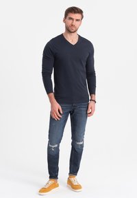 Marineblaues Langarm-V-Ausschnitt-Shirt kombiniert mit hellblauen, verwaschenen Jeans und gelben Sneakers. Schlichtes Design ohne sichtbare Muster oder Akzente.
