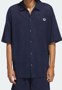 Camicia a maniche corte blu navy realizzata in tessuto a rete traspirante, dotata di un colletto a punta, chiusura a bottoni e un piccolo logo ricamato sul petto.