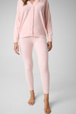 Femme portant un cardigan boutonné rose pâle et un legging ajusté assorti, debout pieds nus sur un sol gris clair.