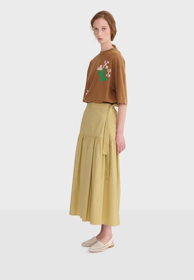 Žena stoji u profilu noseći smeđu majicu s pikseliziranim cvjetnim uzorkom, dugu bež omotnu suknju i svijetlo bež pletene slip-on cipele.