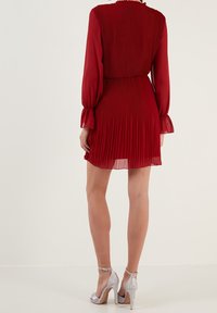 Abito rosso plissettato con maniche lunghe e vita arricciata. Il tessuto è leggero e semitrasparente. Indossato con sandali con tacco alto color argento.