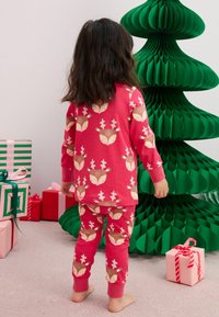 Ensemble de pyjama rouge orné d'un motif de cerfs, avec des manches longues et un pantalon ajusté. En arrière-plan, un sapin de Noël en papier vert et des cadeaux emballés.
