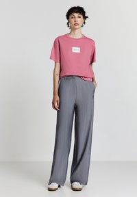 T-shirt rosa a maniche corte con grafica quadrata, abbinato a pantaloni larghi plissettati grigi e sneakers bianche con suole marroni.