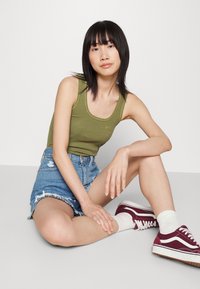 Top sin mangas acanalado verde olivo, pantalones cortos de mezclilla desgastados de tiro alto, zapatillas burdeos, calcetines blancos; pose relajada sentado.