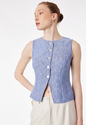 Donna che indossa un top blu senza maniche con bottoni e pantaloni beige chiaro, in posa con le mani dietro la schiena, guarda di lato contro uno sfondo bianco.