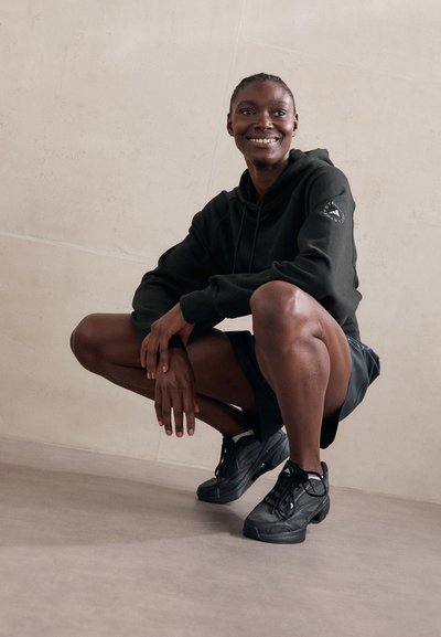 Felpa nera con logo sulla manica, abbinata a pantaloni corti neri e sneakers nere con texture, contro un muro chiaro e liscio.