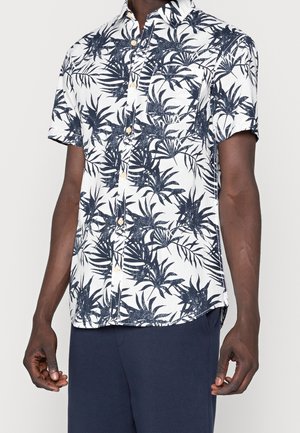 Mand iført hvid kortærmet skjorte med marineblåt tropisk bladmønster og mørkeblå bukser, stående med arme afslappet.