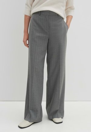 Vrouw die brede grijze pantalon met krijtstreep en witte schoenen draagt, staand met één hand in de zak tegen een effen achtergrond.