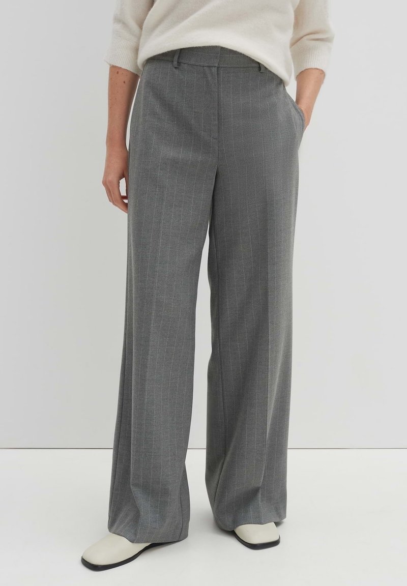 Femme portant un pantalon gris à rayures larges et des chaussures blanches, debout avec une main dans la poche devant un fond uni.
