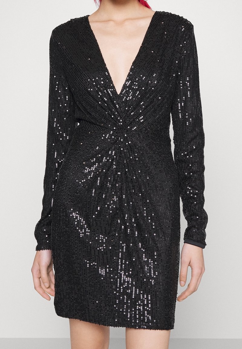 Mini-robe noire à manches longues avec un col en V profond et des sequins sur toute la surface, portée par une personne avec des cheveux roux visibles en haut.