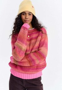 Kobieta z długimi kręconymi włosami, w żółtej czapce typu beanie, różowo-pomarańczowym swetrze w paski, ciemnych spodniach i złotym naszyjniku z zawieszką.