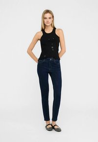 Mustast kaunistustega varrukateta top, kombineeritud tumeliste siniste kitsaste teksadega. Modell kannab musti plätu-sussisid ja seisab neutraalsel taustal.