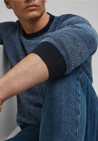 Niebieski sweter w wzory z prążkowanymi ciemnoniebieskimi mankietami, w zestawie z dżinsowymi spodniami. Tekstura ukazuje połączenie gładkich i dzianinowych elementów.