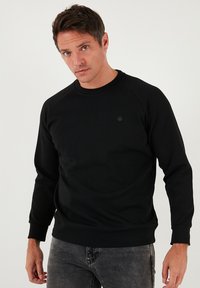 Fekete pulóver hosszú raglán ujjakkal, kerek nyakkivágással és bordázott mandzsettákkal. Finom logó részlet található a mellkason. Puha anyagtextúra.