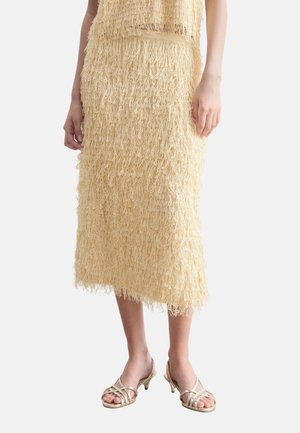Femme portant une jupe midi beige texturée avec des détails en fil bouclé et des sandales dorées à talons bas avec brides, debout sur un fond blanc.