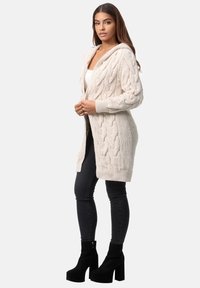 Kabelstickad lång kofta i ljusbeige med huva, med ett texturerat mönster och ribbade muddar, bärs över en vit topp och matchas med svarta jeans.