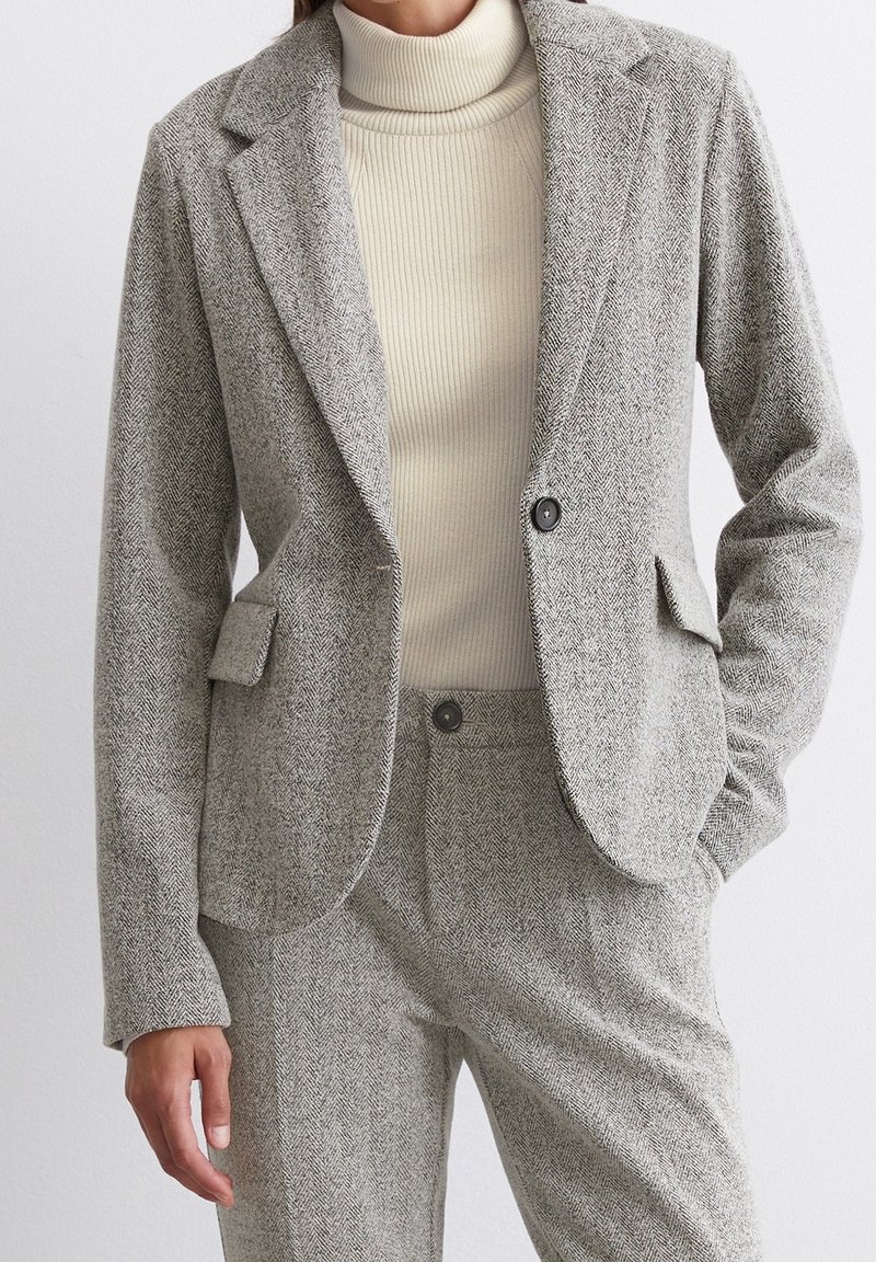 Femme portant un blazer gris à chevrons et un pantalon assorti avec un pull à col roulé côtelé crème. Une main dans la poche.