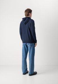 Felpa navy con cappuccio regolabile, maniche lunghe e polsini a coste, indossata con jeans larghi blu chiaro e scarpe nere.