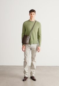 Zöld kötött pulóver, világosszürke farmer, sötétbarna oldaltáska, barna öltönycipő, sima padlón állva, egyszerű háttér előtt.