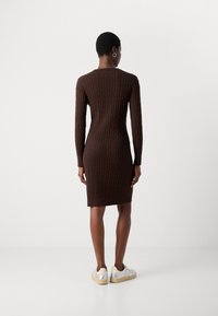 Bruine gebreide jurk met een geribbelde structuur, lange mouwen en een aansluitende silhouet, die net boven de knie eindigt. Casual schoeisel met zilveren accenten.