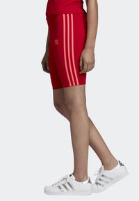 Osoba oblečená v červených šortkách Adidas, které mají tři světlejší červené pruhy po stranách, a v bílých teniskách Adidas se stříbrnými akcenty.