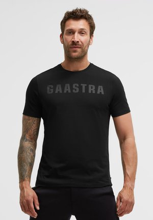 Man with short brown hair and tattooed left arm wearing a black Gaastra t-shirt and dark pants, standing against a plain light background.