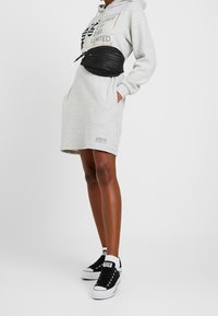 Persona vistiendo un vestido con capucha de color gris claro con texto negro, una bolsa de cintura negra y zapatillas en blanco y negro sobre un fondo blanco.