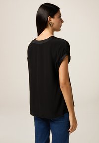 Blusa nera a maniche corte con una trama liscia, dotata di scollatura rotonda e dettagli con spacchi laterali, indossata con jeans blu.