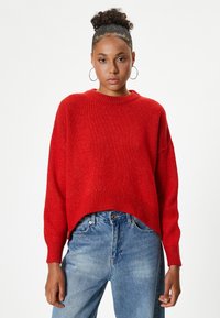 Rode gebreide trui met een ronde hals, verlaagde schouders en ribgebreide zoom. Gecombineerd met hoge taille blauwe denim jeans. Soepele textuur.