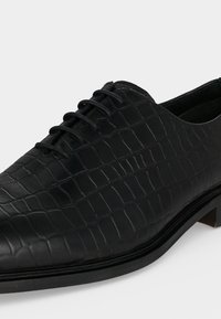 Chaussure de ville en cuir noir avec texture crocodile et laçage fermé sur un petit talon, sur fond uni.