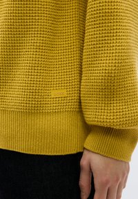 Pull jaune en tricot avec motif texturé, poignets et ourlet inférieur côtelés. Présente une étiquette marron "BOSS" sur le côté gauche.