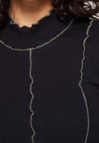 Schwarzer gerippter Rollkragenpullover mit kontrastierenden grünen Rüschennähten entlang des Kragens und der vertikalen Nähte. Weicher Stoff und taillierte Passform.