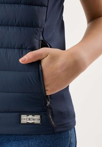Personne avec la main dans la poche zippée d’une doudoune bleu marine, étiquette "Denim Factory" visible, portant un jean bleu sur fond uni.