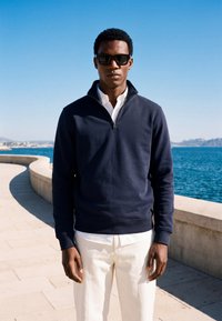 Homme portant des lunettes de soleil foncées, un sweat-shirt bleu marine à demi-fermeture éclair, une chemise blanche et un pantalon blanc, debout sur une promenade au bord de la mer sous un ciel dégagé.