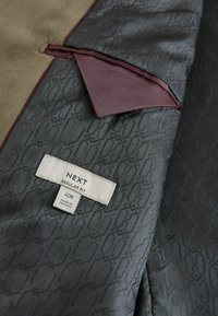Tissu marron et gris avec un motif subtil. Comprend un tissu d'accent maroon, une étiquette avec "NEXT REGULAR FIT 40R" et un design élégant.
