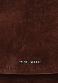 Borsa in suede marrone con logo metallico "Coccinelle", linee pulite e una texture morbida con cuciture visibili lungo il bordo.