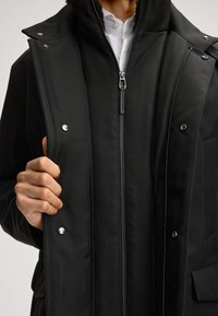 Schwarze Jacke mit einem Reißverschluss an der Vorderseite, Druckknöpfen und einem strukturierten Stoff. Verfügt über einen hohen Kragen sowie sichtbare Reißverschlüsse und Taschen.