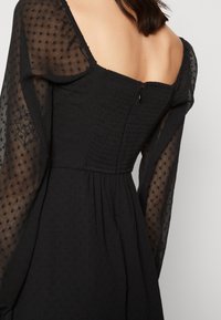 Svart klänning med genomskinliga, puffiga långa ärmar, texturerad bodice med smock och fyrkantig halsringning. Bakom finns en liten dragkedja.