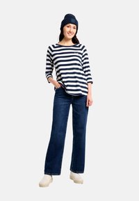 Marineblaue und weiße, gestreifte Langarmshirt, blaue Jeans, weiße Slip-On-Schuhe und eine marineblaue Strickmütze mit Logodetail. Modell posiert mit der Hand in der Tasche.