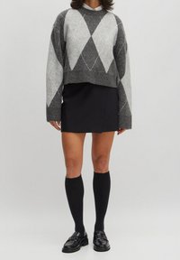 Personne portant un pull à losanges gris et blanc, une mini-jupe noire, des chaussettes noires montant jusqu'aux genoux et des mocassins noirs, se tenant devant un fond uni.