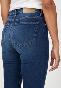 Tmavě modré džíny s vysokým pasem, s kapsami na zadní straně a béžovou koženou etikou na pase. Hladká textura, slim fit.