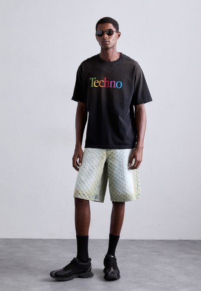 T-shirt en coton noir avec "Techno" en lettres multicolores, associé à un short à motifs vert clair et des baskets noires. Modèle portant des lunettes de soleil.