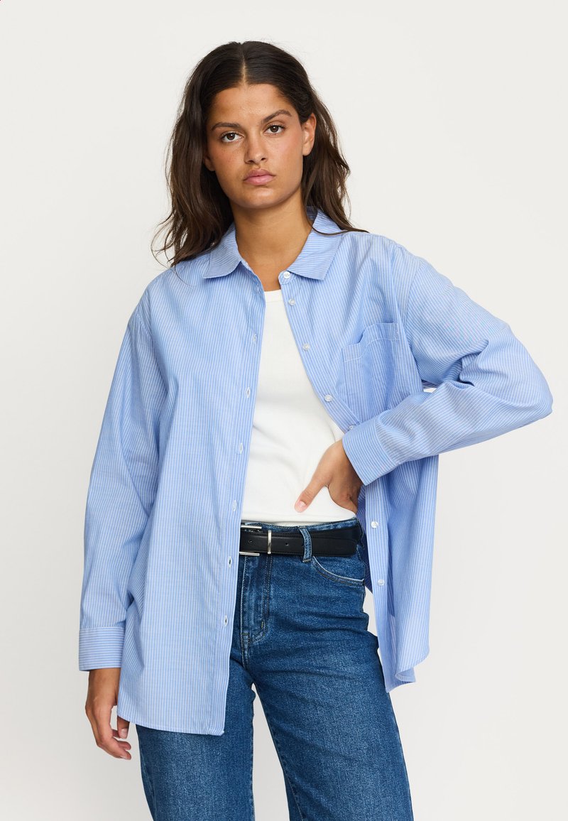 Camicia a righe blu e bianche con colletto, tasca anteriore e maniche lunghe, abbinata a una maglietta bianca e jeans blu.